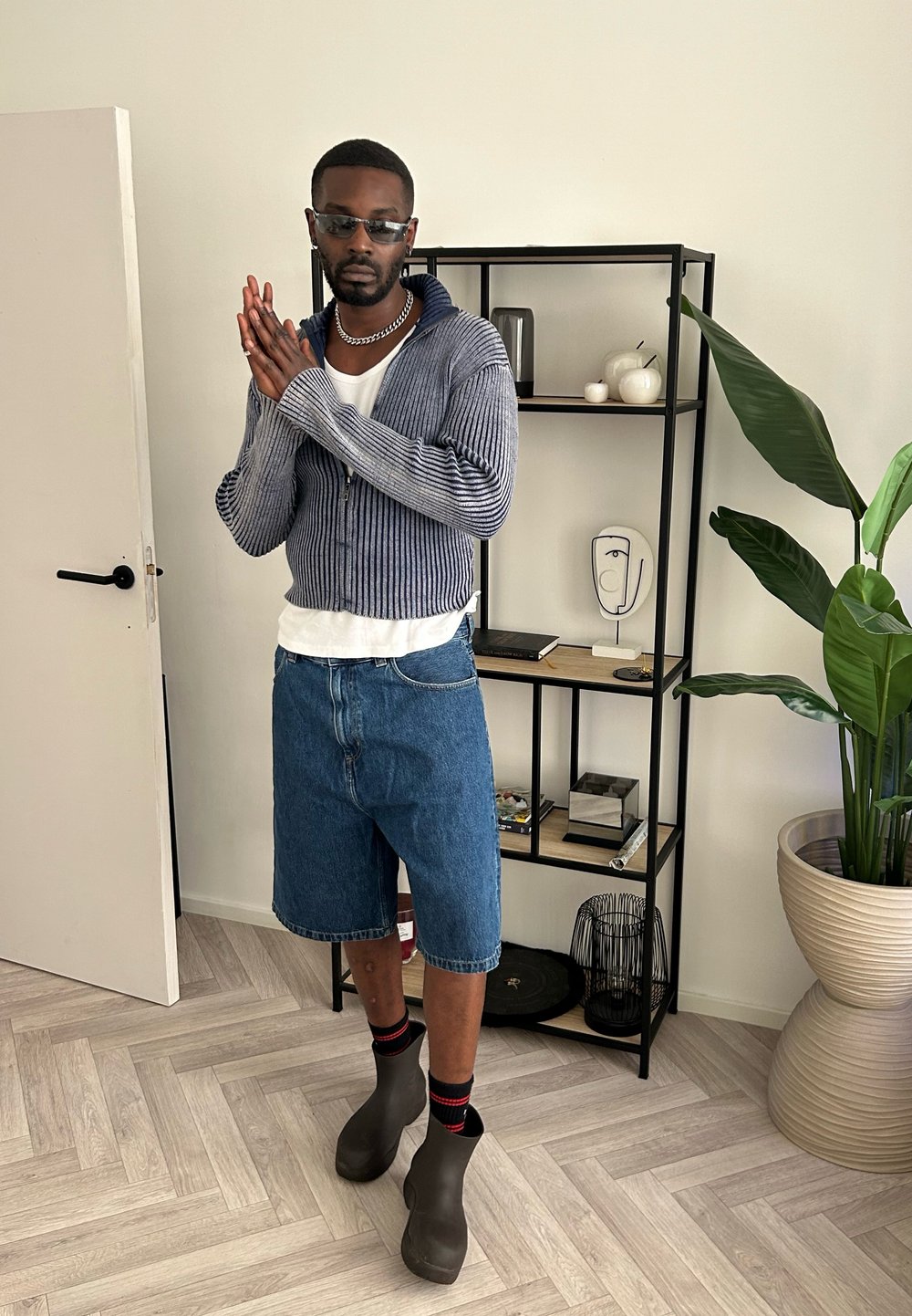 Blåstribet zip-up sweater, hvid t-shirt under, denim shorts, sorte klipklapper, røde og sorte sokker. Indendørs setting med en hylde i baggrunden.