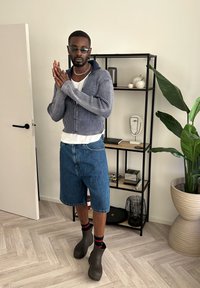 Blåstribet zip-up sweater, hvid t-shirt under, denim shorts, sorte klipklapper, røde og sorte sokker. Indendørs setting med en hylde i baggrunden.