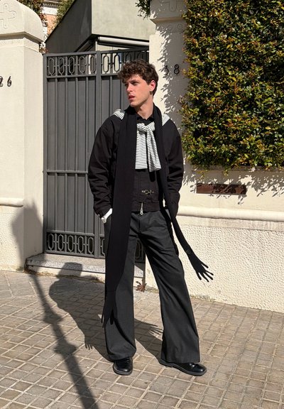 Joven con pantalones anchos negros, chaqueta y larga bufanda con extremos en forma de guantes, de pie en una calle soleada cerca de una puerta y vegetación.