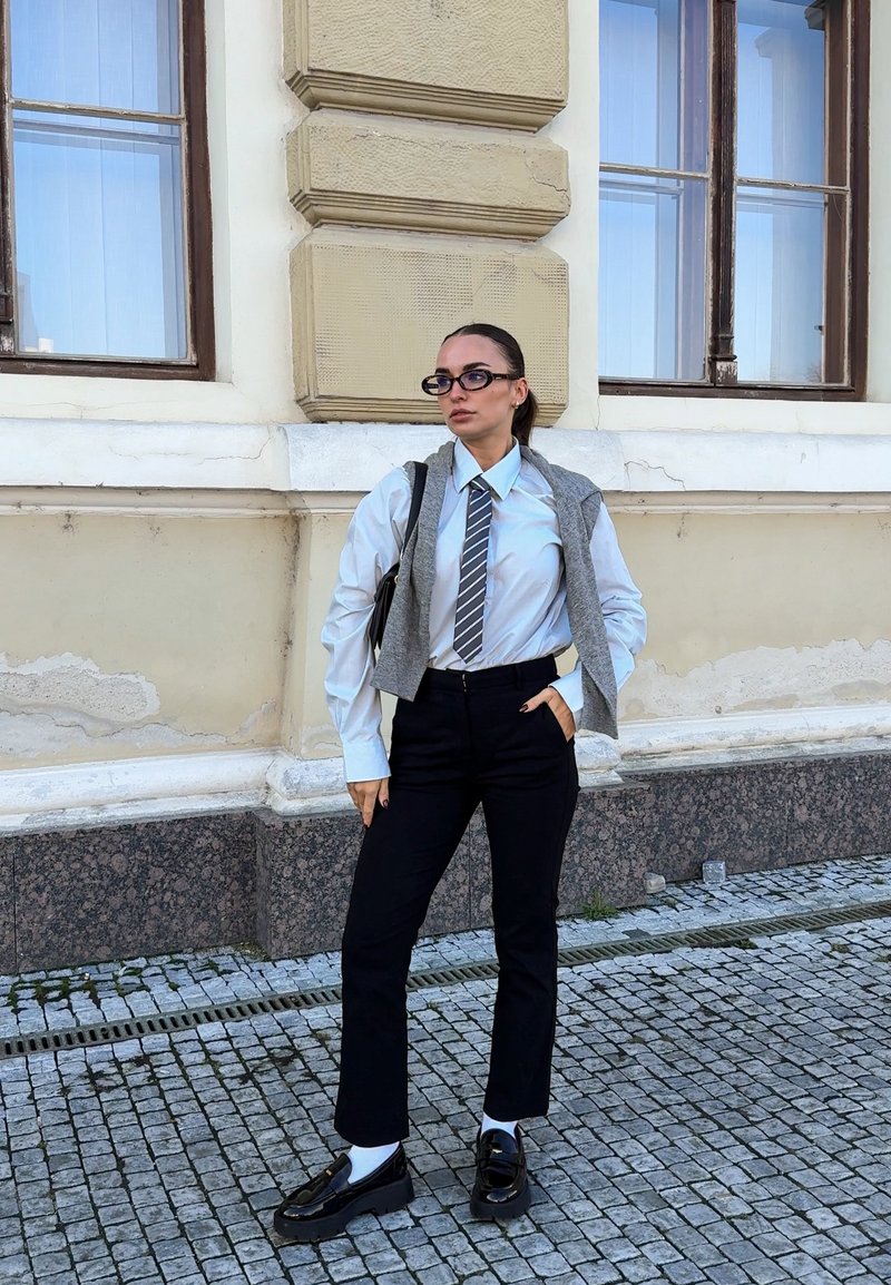 Giovane donna con occhiali, camicia bianca, cravatta a righe, maglione grigio sulle spalle, pantaloni neri e mocassini che si trova su una strada lastricata.