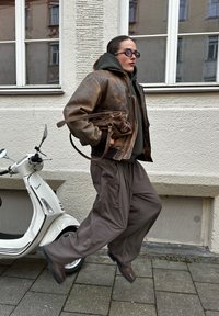 Barna bőrkabát zöld kapucnis pulóver felett, laza szürke nadrág, barna csizmák; barna táskát tartva, és elhalad egy fehér robogó mellett.