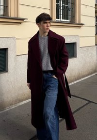 Manteau en laine bordeaux porté sur un pull gris à torsades et un jean large bleu, debout sur un trottoir en ville avec un mur beige en arrière-plan.