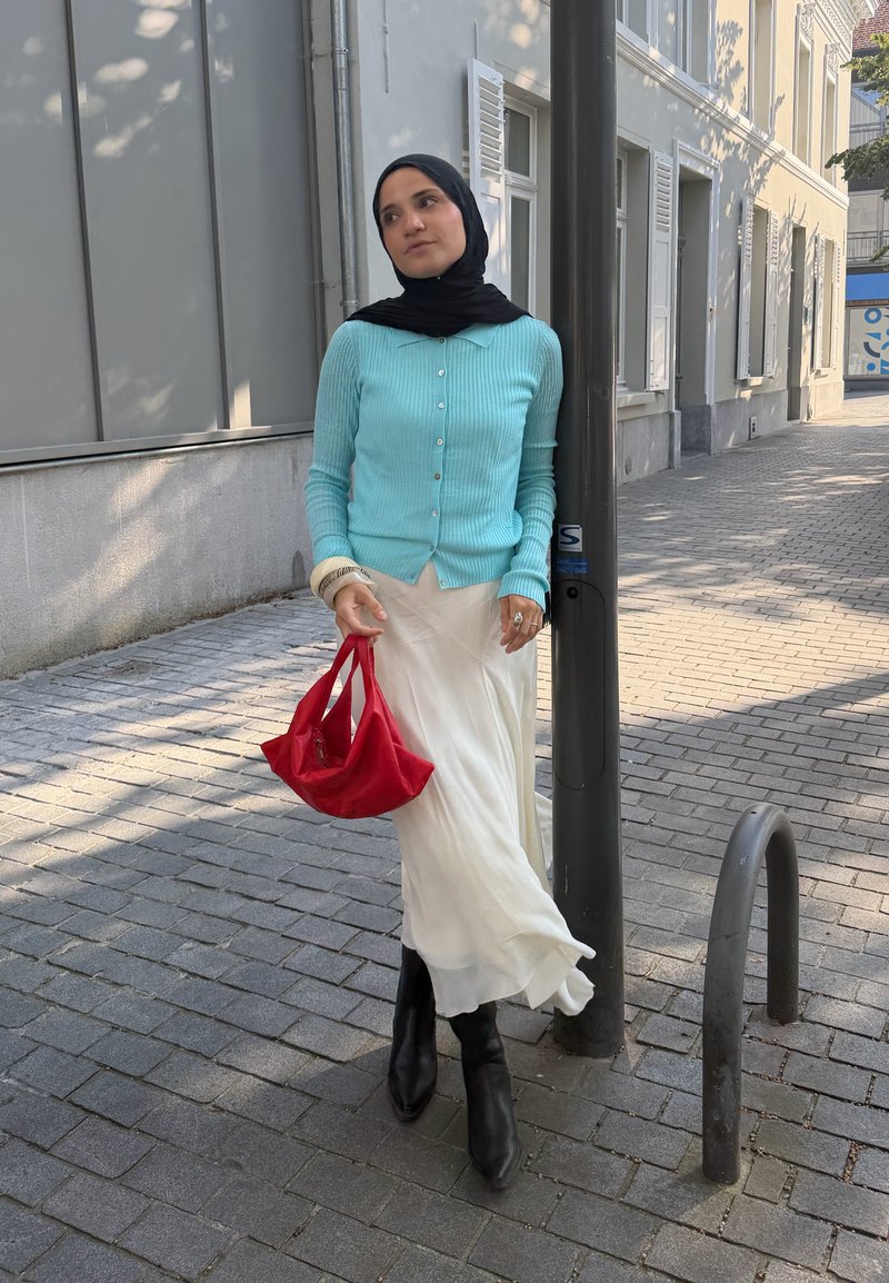 Woman wearing black headscarf, blue cardigan, white skirt, and black boots standing on a cobblestone street holding a red bag.