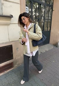 Beige jacket, pink striped shirt, grey trousers, and light shoes. Black handbag with a soft texture. Strolling along a cobbled street.