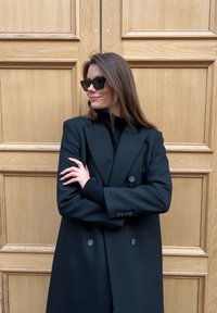 Black double-breasted coat with wide lapels, textured fabric, and dark knit sweater underneath. Model wearing angular black sunglasses.