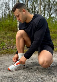 Skriešanas apavi ar baltu sieta augšdaļu, oranžiem akcentiem, melniem auklām un unikālu mīkstinātu zoli, kas fiksēti dabiskā fonā.