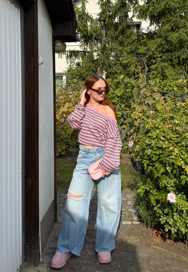 Joven mujer con top rosa a rayas sin hombros, vaqueros anchos desgarrados y zapatos rosas, de pie en un camino cerca de vegetación y un edificio.