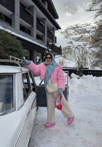 Personne en manteau rose et écharpe bleue fixant un sapin de Noël sur le toit d'une voiture ancienne dans une station de montagne enneigée.