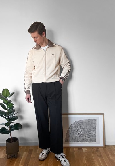 Chaqueta de chándal beige con cuello marrón y tres rayas, pantalones de pierna ancha negros y zapatillas blancas. Configuración interior con una planta y una obra de arte.