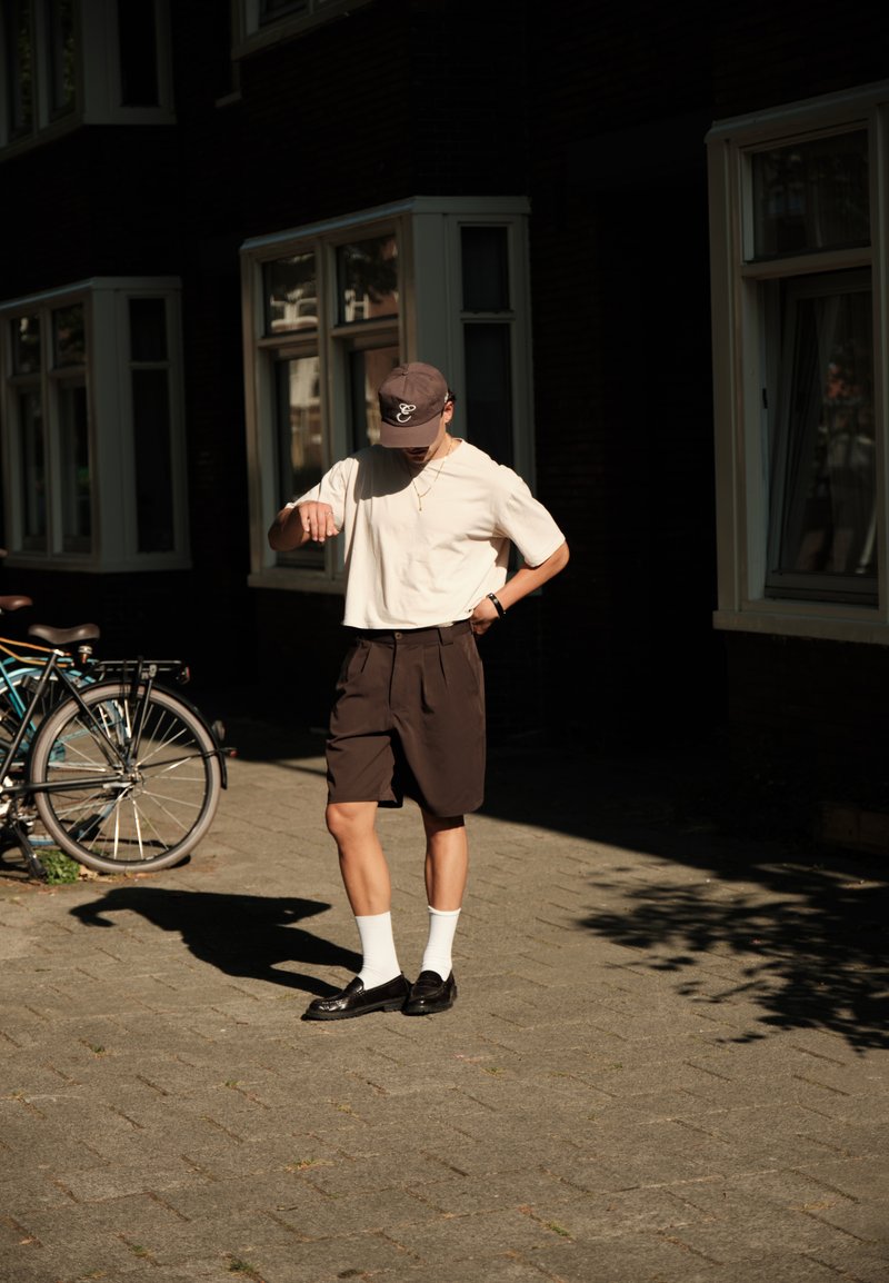 Person wearing a cap, white shirt, dark shorts, white socks, and black shoes standing on a sunlit sidewalk near bicycles and windows.