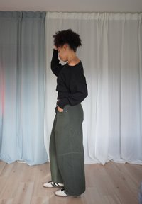 Schwarzer, kurz geschnittener Pullover mit Off-Shoulder-Design, kombiniert mit weiten, olivgrünen Hosen. Das Model trägt beigefarbene Sneakers, beleuchtet vom Hintergrund durch einen Vorhang.
