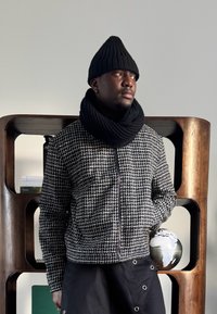 Homme portant un bonnet en laine noir, une écharpe noire et une veste à carreaux noir et blanc, debout devant une étagère en bois avec un globe terrestre.