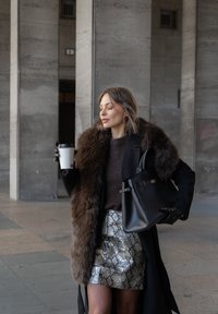 Čierny kabát s kožušinovým golierom, hnedý sveter, strieborná sukňa z hadej kože, držiaca šálku kávy a čiernu kabelku s elegantným dizajnom.