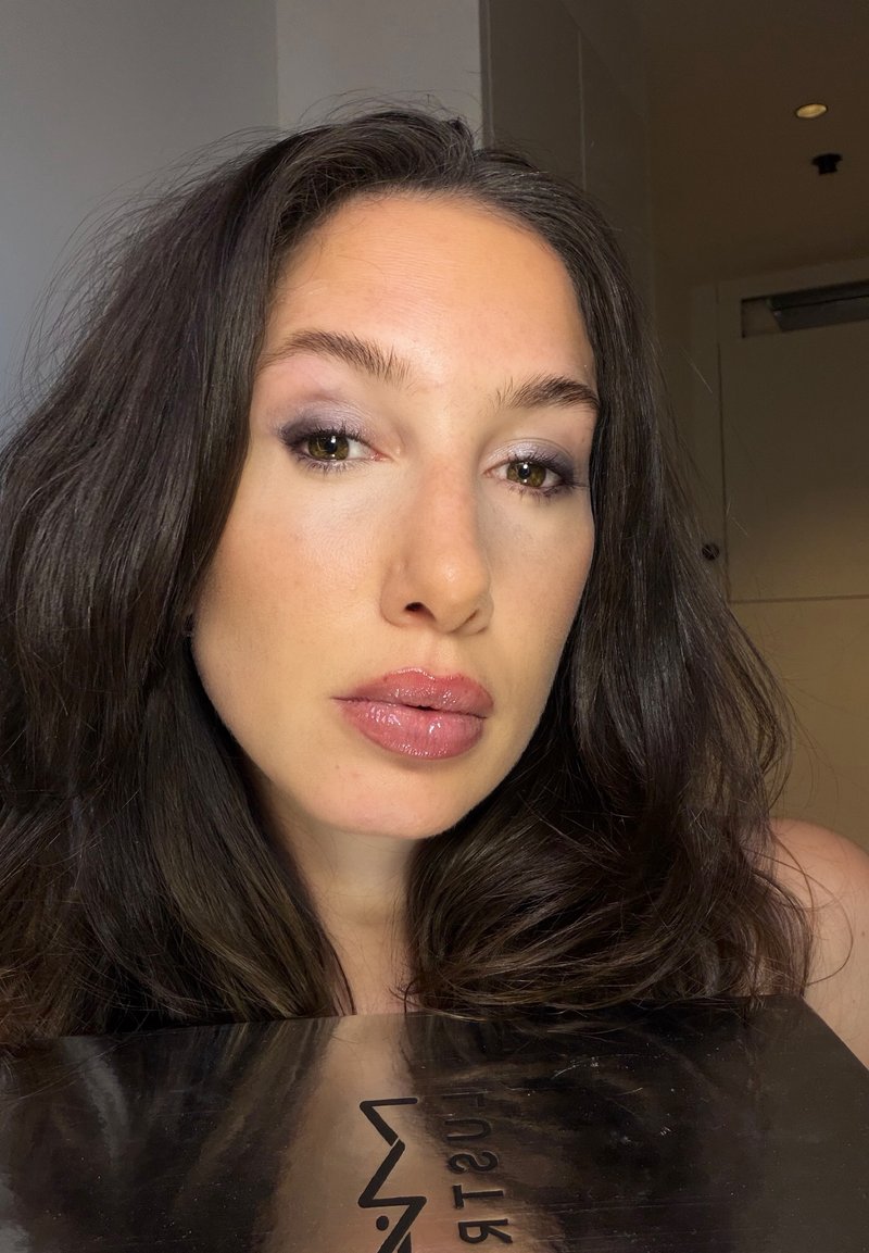 Femme aux longs cheveux foncés et aux lèvres roses brillantes regardant la caméra, tenant une surface noire réfléchissante avec un texte partiellement visible.