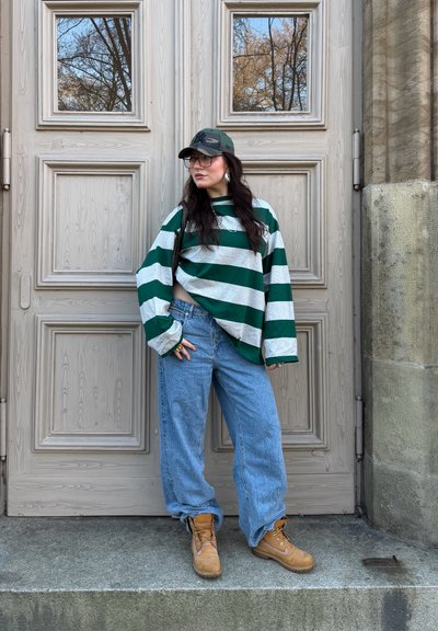 Sudadera grande de rayas verdes y grises, vaqueros holgados azul claro, botas color canela y gorra de camuflaje, delante de puertas de madera con ventanas.