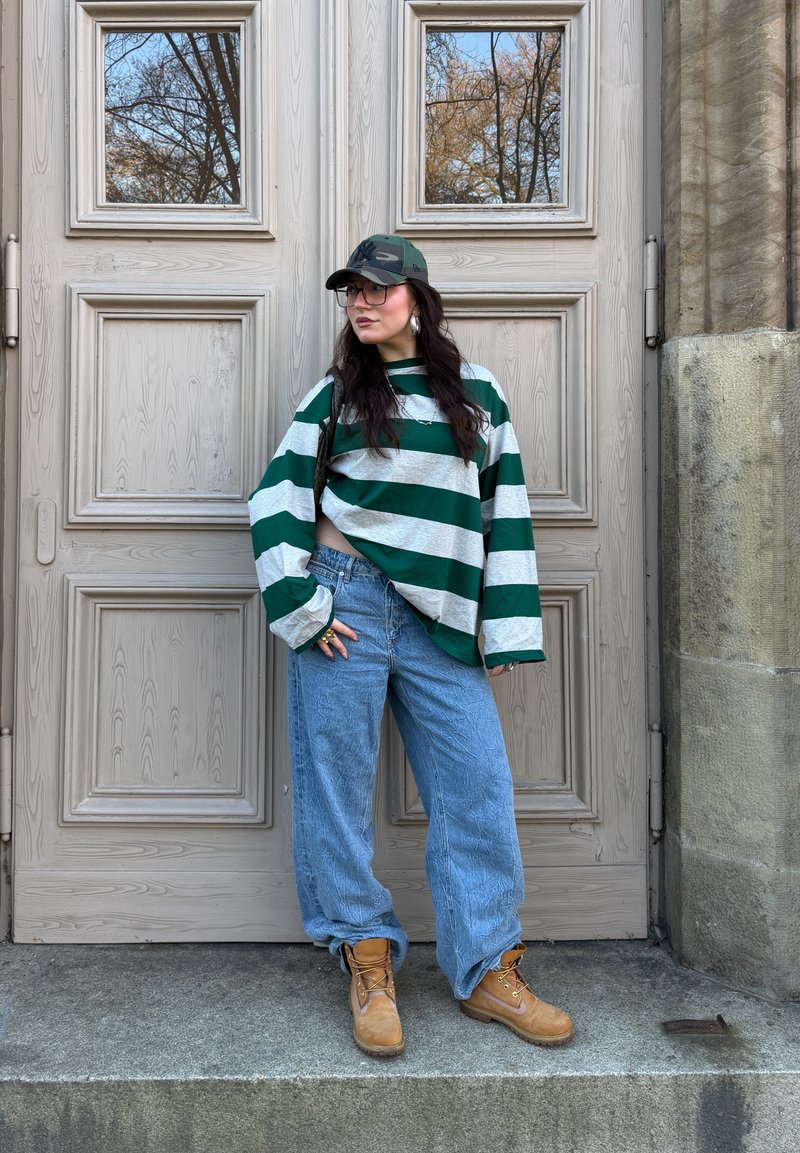 Sweat-shirt surdimensionné rayé vert et gris, jean ample bleu clair, bottes couleur tan et casquette camouflage, devant des portes en bois avec fenêtres.