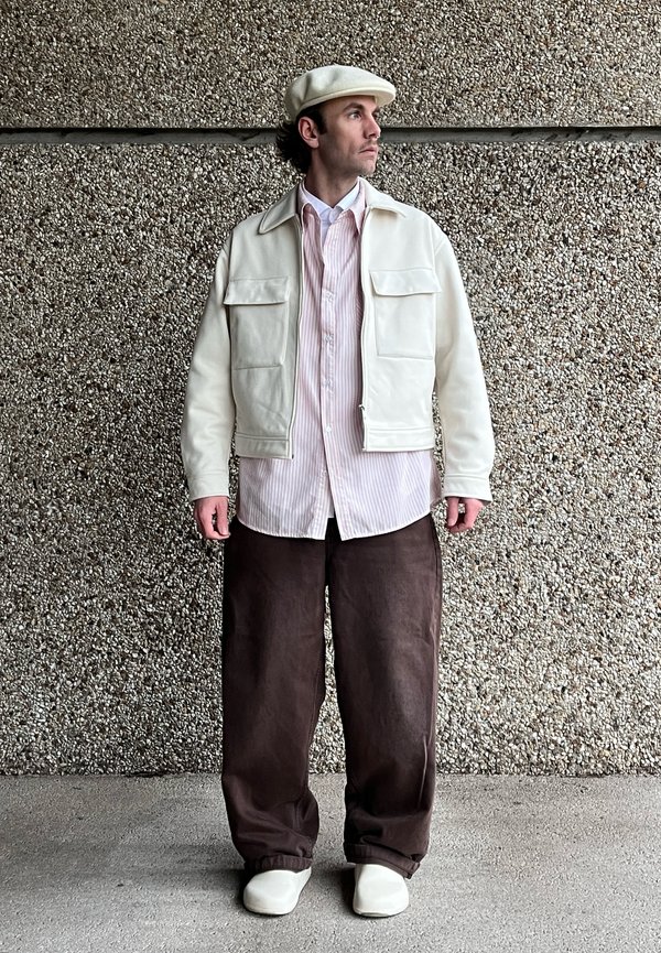 Chaqueta de cuero crema con dos bolsillos delanteros sobre una camisa a rayas rosa claro y blanca, combinada con pantalones sueltos marrones y zapatillas blancas.