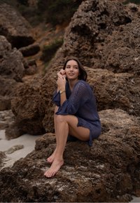 Cover-up de plajă tricotat albastru cu un design lejer și țesătură texturată, purtat peste picioarele goale, așezat pe un teren stâncos lângă țărm.