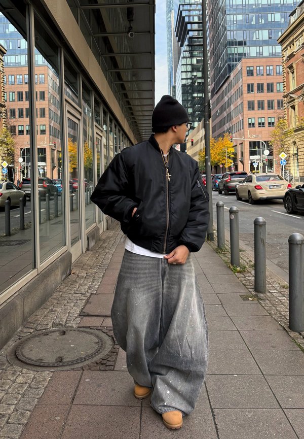 Personne portant un bonnet noir, une veste noire et un pantalon large gris marchant sur un trottoir urbain avec un bâtiment en verre et des voitures en arrière-plan.