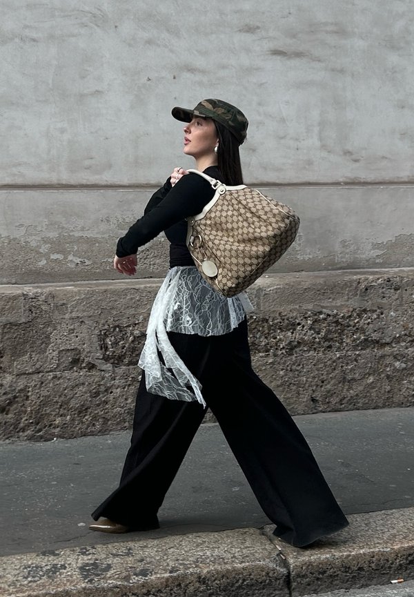 Žena v maskáčovej čiapke, čiernom tričku, širokých čiernych nohaviciach a čipkovanom opasku kráča popri kamennom múre a nesie veľkú vzorovanú béžovú kabelku.