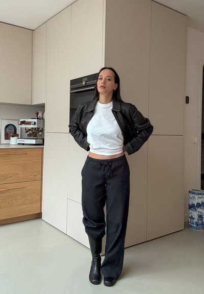 Chaqueta bomber de cuero negro sobre una camiseta blanca corta, combinada con pantalones negros sueltos y botines negros. Fondo de cocina minimalista.