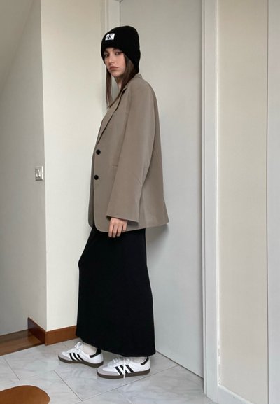 Blazer beige de gran tamaño con botones negros, combinado con una falda larga negra y zapatillas blancas con rayas negras, frente a un fondo simple.