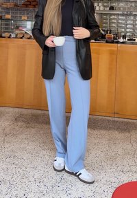 A woman standing in a café wearing light blue trousers, a black leather jacket, and white trainers, holding a white coffee cup.