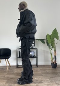 Czarna kurtka bomber z kołnierzem z futerka ekologicznego, zestawiona z przetartymi czarnymi spodniami cargo oraz masywnymi czarnymi botkami na platformie. Wnętrze z roślinami.