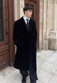 Long manteau noir en velours à double boutonnage avec revers à pointe, porté par-dessus un costume noir et une cravate. La silhouette se tient devant un décor en pierre historique.