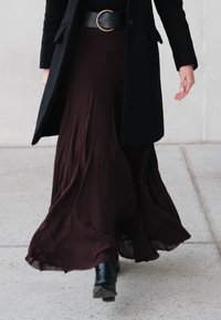 Longue jupe fluide bordeaux avec une finition texturée, associée à un manteau noir et des bottines, avec une boucle de ceinture circulaire.