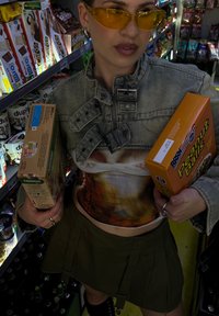 Traper jakna s grafičkim dizajnom, maslinasto zelena plisirana suknja, žute naočale, drži kutiju žitarica i kutiju grickalica u prolazu trgovine.