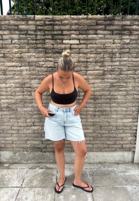 Vaaleansiniset farkkushortsit, joissa on rispaantuneet reunat, yhdistetty mustaan lyhytpaita-toppiin. Mustat varvassandaalit, joita käytetään betonipinnalla, taustalla kiviseinä.