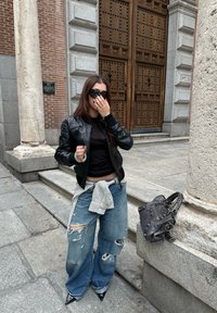 Črna očesana usnjena jakna čez črno majico, kombinirana z raztrganimi širokimi modrimi kavbojkami, zavezanimi s karirasto krpo, ter z ostrimi črnimi petami.