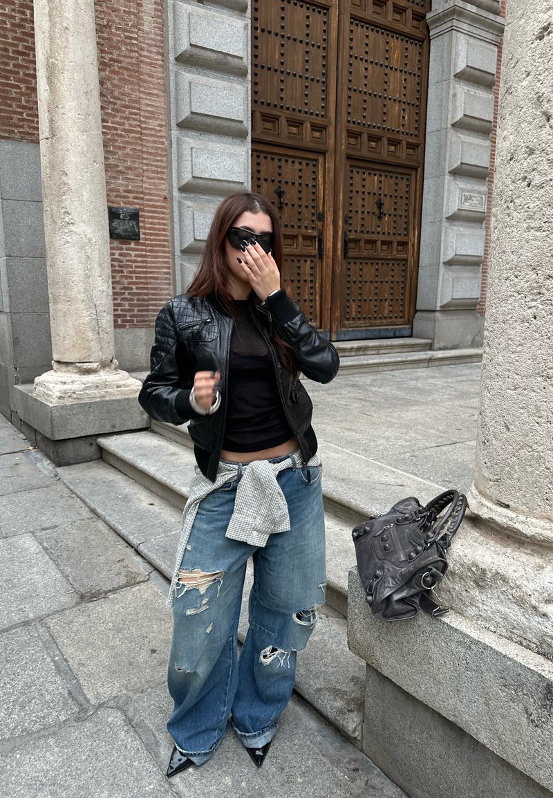 Schwarze gesteppte Lederjacke über einem schwarzen Oberteil, kombiniert mit zerrissenen, weiten blauen Jeans, die mit einem karierten Tuch gebunden sind, und spitzen schwarzen High Heels.