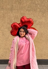 Femme en manteau rose et sweat-shirt tenant deux ballons rouges en forme de c œur devant un mur beige texturé.