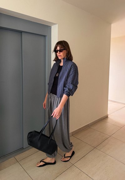 Mujer con gafas de sol, chaqueta a rayas azul marino, pantalones grises holgados y chanclas negras, sujetando un bolso negro cerca de unas puertas de ascensor grises.