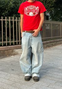 T-shirt en coton rouge avec motif imprimé, jean large en denim bleu clair, ceinture en cuir marron, et sabots noirs sur une surface pavée.