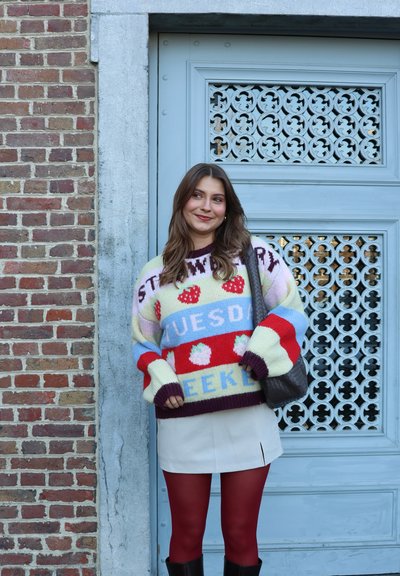 Mujer joven con suéter colorido con estampado de fresas y falda blanca, de pie sonriendo junto a una puerta azul ornamentada y una pared de ladrillos.