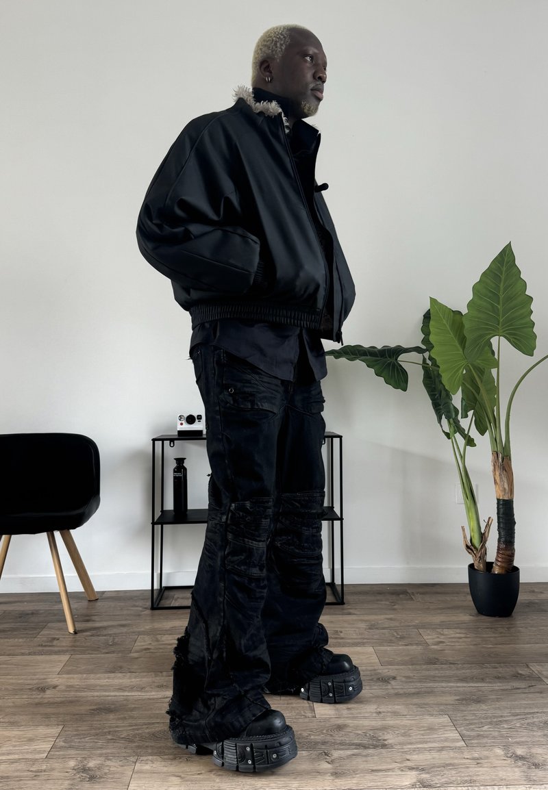 Black bomber jacket with fur collar, black textured cargo pants, and chunky black footwear. Indoor setting with green plant and furniture.