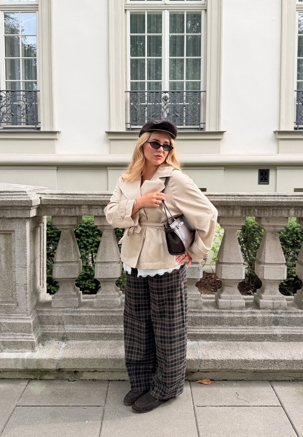 Femme blonde portant un manteau beige, un pantalon large à carreaux, un béret noir, des lunettes de soleil et un sac sombre, debout près d'une rambarde en pierre devant un bâtiment avec fenêtres.
