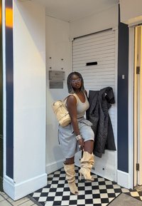 Sieviete, tērpusies pelēkā kleitā, ar bēša zābakiem un mugursomu, pozē uz melnbalta šaha dēļa grīdas pie baltām un zilām durvīm.