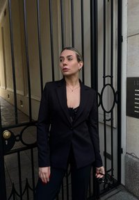 Femme en blazer noir et haut en dentelle appuyée contre une grille en fer noir, tenant des lunettes de soleil, regardant sur le côté dans un couloir urbain étroit.