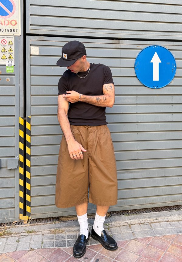 Mann mit schwarzer Kappe, schwarzem T-Shirt, brauner weiter Shorts, weißen Socken und schwarzen Loafers steht an einer Metallwand mit einem Pfeil nach oben Zeichen.