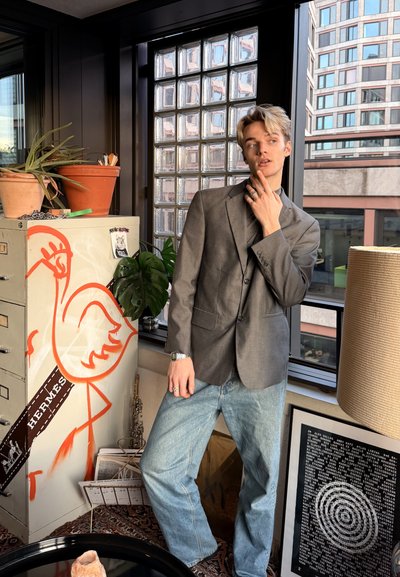 Joven con blazer gris y vaqueros posa en interiores junto a un archivador pintado, plantas, obras de arte enmarcadas y una gran ventana con vista a la ciudad.
