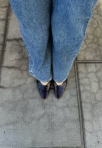 Chaussures marron à bouts pointus associées à un jean en denim bleu clair à jambes larges, présentant un ourlet effiloché, sur une surface grise texturée.