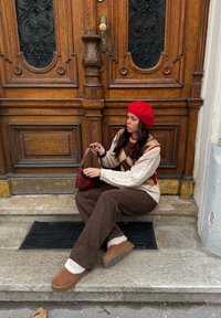 Béret rouge, pull crème avec motifs rouges et beiges, pantalon large marron, et chaussons beiges. Tient un petit sac à main rouge, assise sur des marches.