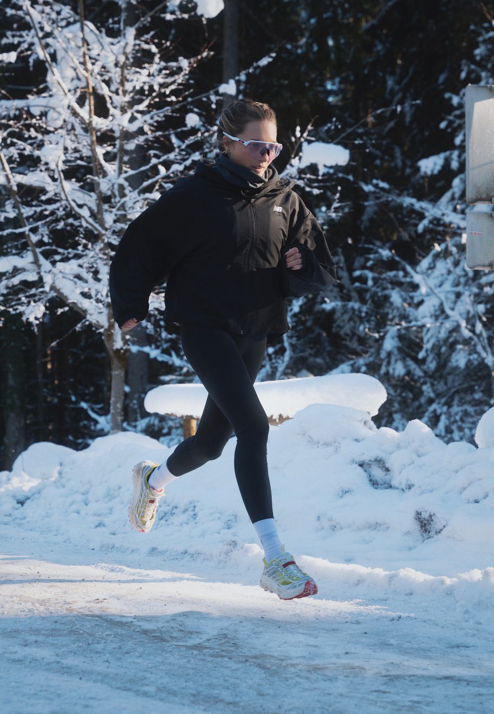 Black ski jacket, black leggings, and vibrant running shoes with a mesh upper. Snow-covered forest backdrop featuring white trees and a blanket of snow on the ground.