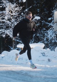 Black ski jacket, black leggings, and vibrant running shoes with a mesh upper. Snow-covered forest backdrop featuring white trees and a blanket of snow on the ground.