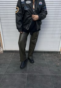 Črna usnjena bomber jakna s našitki, črna majica, olivno zelene hlače iz umetnega usnja in črne slip-on čevlje z teksturiranimi podplati.