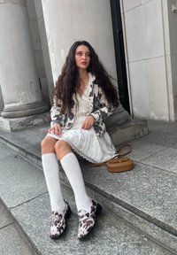 Woman seated on stone steps, wearing a white dress with lace trim, a patterned grey and white jacket, knee-high white socks, and leopard print loafers.
